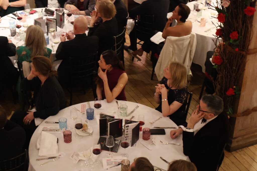 Guests listening to a speech, sat at a round dining table at a black tie Create annual Gala.