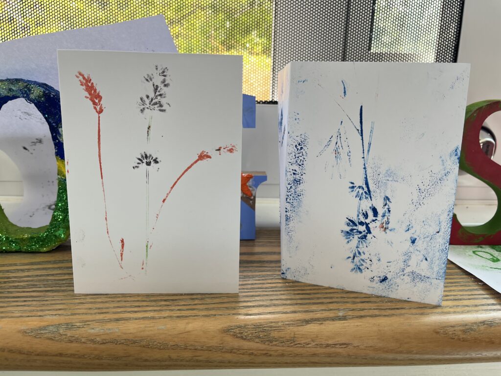 Two pieces of white paper folded into cards, with outlines of plants that have been printed on using blue orange and black ink, taken at a creative:tandem project.